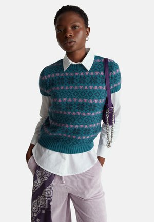 Maglione lavorato a maglia color verde smeraldo con motivi geometrici rosa, indossato sopra una camicia bianca con colletto. Pantaloni viola con motivo a fantasia, accompagnati da una borsa laterale con catena argentata.