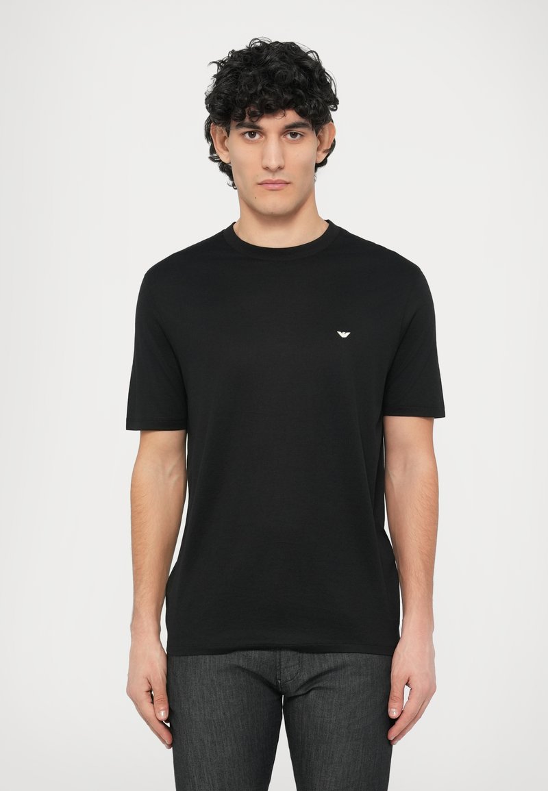 Juoda medvilninė marškinėliai su apvalia iškirpte, trumpomis rankovėmis ir mažu emblematu kairėje krūtinės pusėje. Minkšta tekstūra, laisvas siluetas.