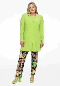 Groene lange mouwen knoopjack over een bijpassende top, gecombineerd met gestreepte broek met kleurrijke bladdessins en groene hakken.