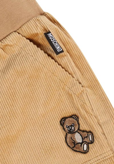 Tessuto di velluto a coste color beige, con una texture a righe, che presenta un ricamo di un orsetto marrone e un'etichetta nera Moschino sulla cintura.