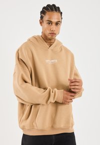 Joven con cabello trenzado ajustando la manga de una sudadera beige con el texto "OFF LIMITS" en el frente, vistiendo pantalones oscuros frente a un fondo simple.