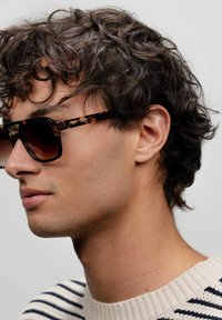 Occhiali da sole tartarugati con lenti scure, dotati di una montatura rettangolare, indossati da una persona con capelli ricci e un maglione a righe.