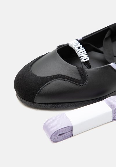 Ballerine en cuir noir et daim avec bride élastique ornée de perles blanches formant le mot "Moschino", à côté d'un ruban violet clair enroulé.