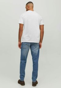 Witte T-shirt met korte mouwen, blauwe denim jeans met lichte vervaging en bruine schoenen, van achteren bekeken tegen een neutrale achtergrond.