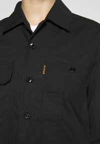 Giacca nera in tessuto testurizzato con colletto classico, chiusura a bottoni, due tasche sul petto e un sottile logo giallo.