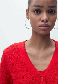 Rode gebreide cardigan met V-hals, voorzien van ruitpatronen, knoopsluiting en textiel met structuur. Model draagt oorbellen met een ring.