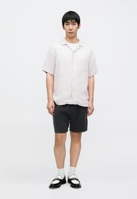 Chemise rose clair à manches courtes avec une poche, short noir, chaussettes blanches et chaussures noires avec détails décoratifs. Le modèle pose devant un fond blanc.