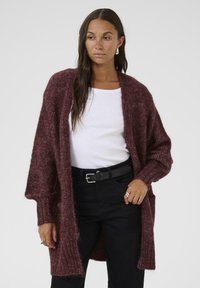 Burgundy knit cardigan with an open front, oversized sleeves, and a textured finish. Worn over a white shirt and black belt.