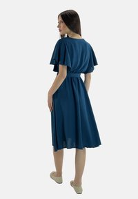 Türkise knielange Kleid mit schwingenden Ärmeln, tailliertem Bund und fließendem Rock. Glatte Textur, leichter Stoff. Legere Schuhe.
