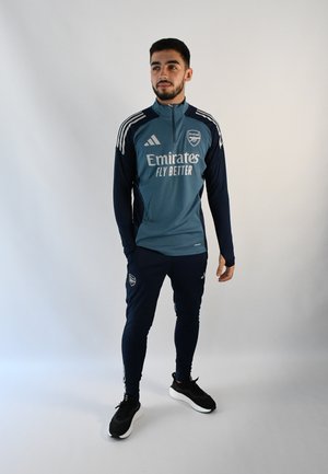 Chaqueta de entrenamiento azul y verde azulado del Arsenal con cremallera, mangas largas y rayas blancas de Adidas. Combinada con pantalones azul marino y zapatillas deportivas negras.