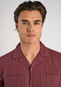 Camicia corta maroon con tessuto testurizzato, due tasche al fronte e chiusura a bottoni. Presenta un colletto classico e una vestibilità comoda.