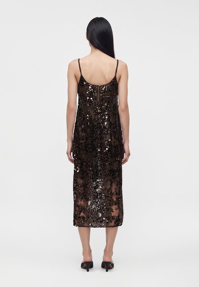 Femme aux longs cheveux noirs de dos, portant une robe midi noire à sequins avec fines bretelles et des chaussures à talons noires sur fond blanc.