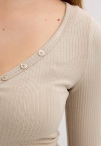 Top beige de canalé con un escote en forma de V profundo y cuatro botones pequeños a lo largo del cuello. La tela tiene un acabado suave y texturizado.