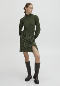 Zeleni pleteni pulover s turtleneckom v kombinaciji z zeleno in črno prugasto mini krilo, ki ima stranski razporek in gumbne detajle. Rjavi škornji do kolen.