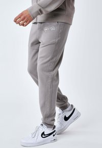 Pantalon de jogging gris en tissu doux, avec un logo latéral en impression en relief. Associé à des baskets blanches avec des accents noirs.
