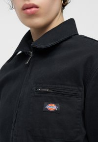 Κοντινό πλάνο ατόμου που φοράει μαύρο τζάκετ με φερμουάρ, με τσέπη στο στήθος και πολύχρωμο λογότυπο Dickies.