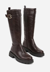Bottes en cuir marron jusqu'au genou avec une finition texturée, dotées d'une fermeture éclair sur le côté et d'une accentuation par une boucle décorative sur le haut de la tige. Semelle à crampons épais.