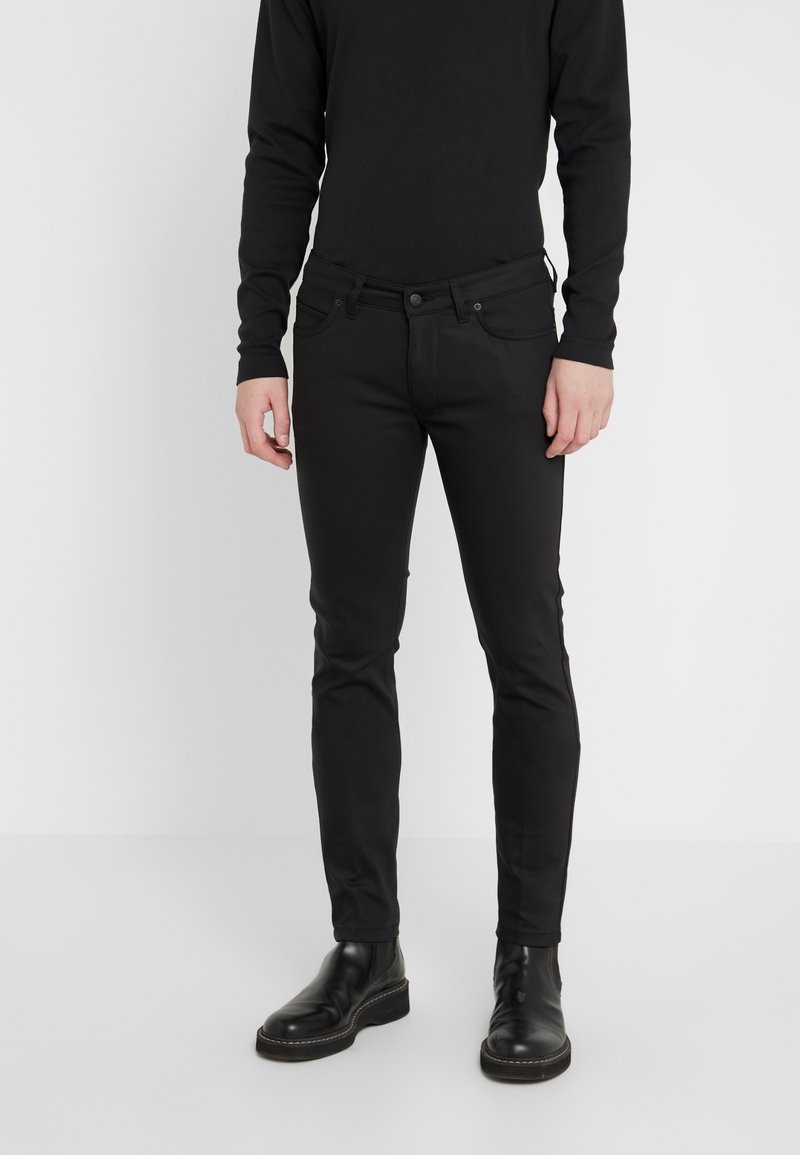 Černé slim-fit džíny s hladkou texturou, pětikapsovým designem, kovovým zapínáním a nenápadnými švy, doplněné o černé boty.
