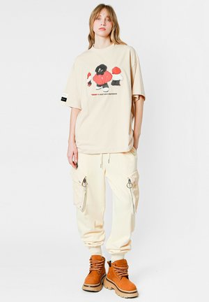 Camiseta oversized beige con un diseño gráfico colorido, combinada con pantalones cargo en color crema que cuentan con cremalleras y bolsillos, y botas de tobillo marrones.