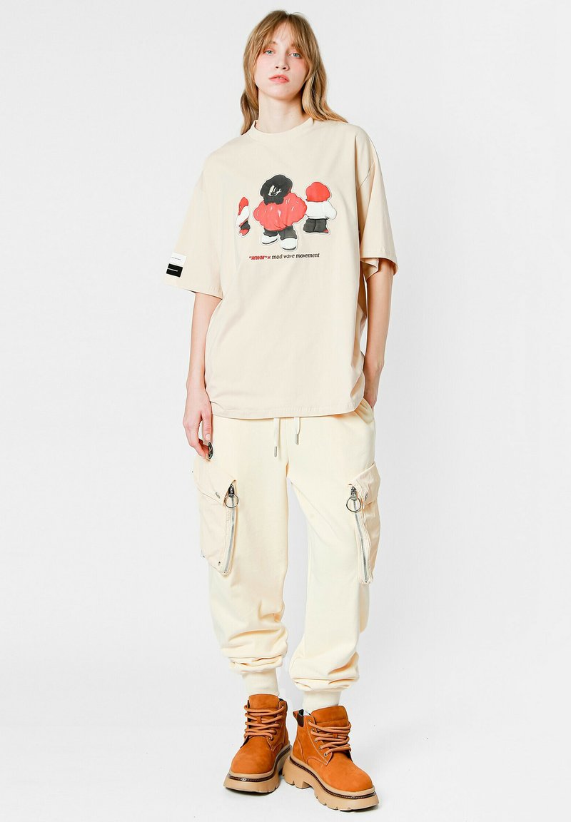 Camiseta oversized beige con un diseño gráfico colorido, combinada con pantalones cargo en color crema que cuentan con cremalleras y bolsillos, y botas de tobillo marrones.