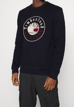 Mężczyzna ubrany w czarną, długorękawową bluzę z okrągłym logo Timberland Hilfiger w beżowym, białym i czerwonym kolorze na piersi.