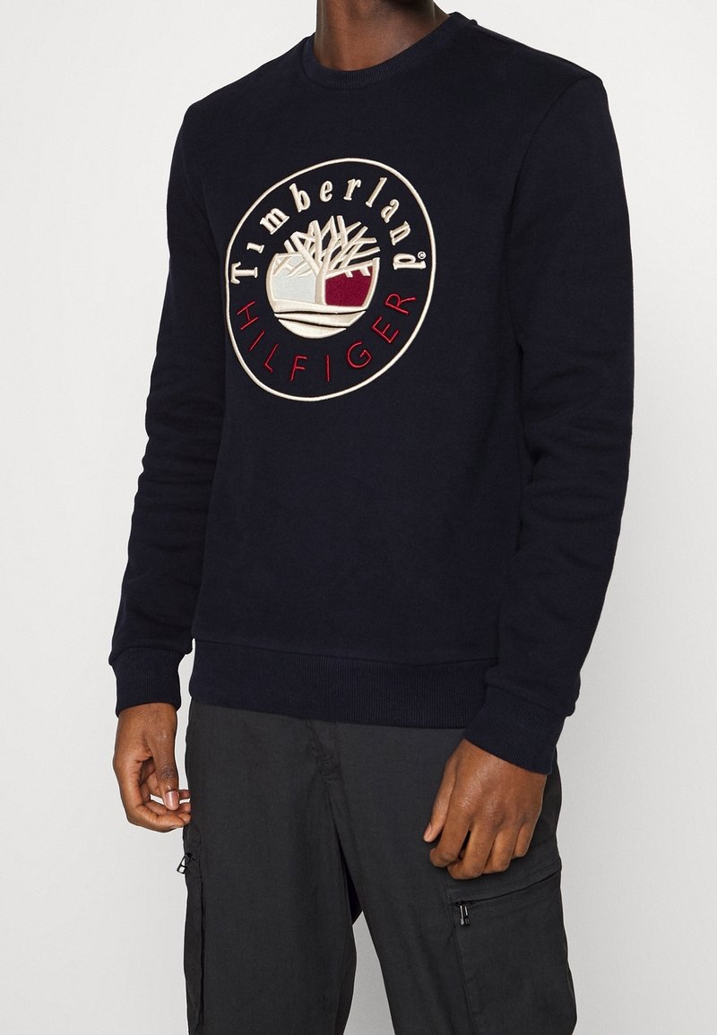 Homme portant un sweat-shirt noir à manches longues avec un logo circulaire Timberland Hilfiger en beige, blanc et rouge sur la poitrine.