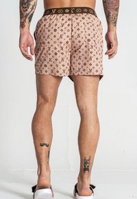 Pantaloni corti marrone chiaro con un design a motivi e loghi. Vita nera con dettagli del logo ripetuti. Gambe scoperte e tatuaggi visibili.