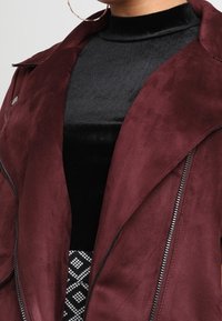 Giacca in finta pelle scamosciata color bordeaux con rever a lancia, chiusura con zip e dettagli in argento. Indossata sopra una maglia in velluto nero e una gonna con motivo.