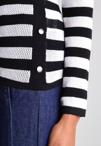 Suéter de punto a rayas en blanco y negro con textura de malla y puños acanalados suaves, que presenta botones metálicos en el lateral.