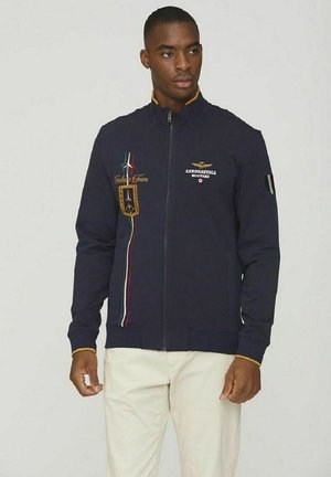 Giacca zip-up blu navy con colletto alto, caratterizzata da strisce multicolori, loghi ricamati e dettagli con toppe. Realizzata in un tessuto morbido, ha un design aderente.