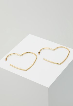Boucles d'oreilles créoles en métal doré en forme de cœurs posées sur un cube blanc sur fond gris clair.