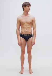 Μαύρα ανδρικά μαγιό με χρυσό μοτίβο ελληνικού κλειδιού στη μέση, με εφαρμοστή σχεδίαση και λεία υφή. Το μοντέλο στέκεται ξυπόλητο.