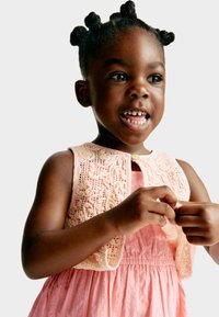 Fille portant une robe rose avec un gilet en dentelle crochetée. Le gilet présente des motifs complexes et une fermeture à bouton au niveau du cou.