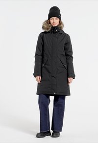 Sort parka med faux fur-besætning på hætten, lynlås lukning og sidelommer. Parret med mørke brede bukser og sorte støvler.