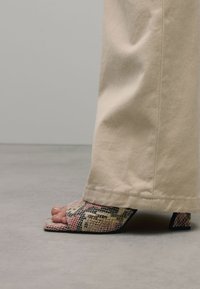 Des sandales à talons avec un motif de serpent texturé et coloré, une conception à bout ouvert et un talon carré, associées à un pantalon de couleur crème.