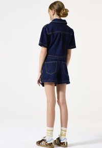 Denim romper met korte mouwen en short, donkerblauw, met zakdetails en contraststiksels. Gecombineerd met gestreepte sneakers en sokken.