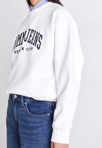 Sudadera blanca con "TOMMYJEANS" y "NEW YORK CITY" en letras marinas. Cuenta con un cuello redondo y puños acanalados. Combinada con unos vaqueros de mezclilla azules.