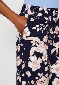 Granatowe kwiatowe spodnie z elastycznym paskiem, z jasnoróżowymi kwiatami i zielonymi liśćmi. Widoczna kieszeń boczna; materiał wydaje się gładki i lekki.