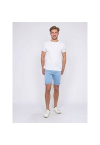 T-shirt blanc à manches courtes, shorts bleu clair avec des revers, et baskets blanches, se tenant devant un fond uni.