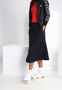 Černá bundě na zip, červené tričko, námořnická midi sukně, bílé platformové sandály s korkovými detaily, nošeno s bílými podkolenkami; minimalistický styl.