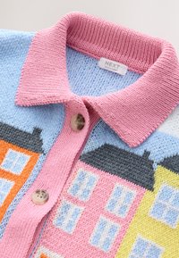 Pletený cardigan s růžovým límcem a knoflíky, s barevnými vzory domů v oranžové, růžové, žluté a modré na světle modrém pozadí.