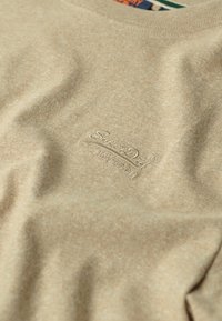 Beige fabric with subtle embroidered SuperDry logo and Japanese text on soft, textured cotton material.