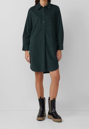 Femme portant une robe-chemise à manches longues vert foncé et des bottines noires épaisses avec des semelles beiges, debout devant un fond uni.