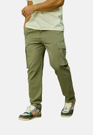 Homme portant un pantalon cargo vert olive et des baskets multicolores, la main dans la poche, debout devant un fond uni.