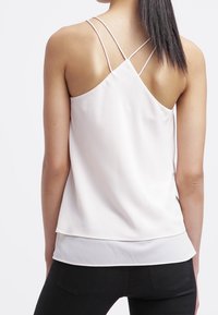 Jasnoróżowy top bez rękawów z niskim plecami, z cienkimi krzyżującymi się ramiączkami. Wykonany z gładkiego materiału, z warstwowym dołem na czarnych spodniach.