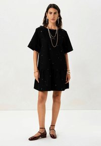 Schwarzes A-Linien-Kleid mit kurzen Ärmeln, verziert mit buntem floralen Stickerei und einem stufigen Saum. Kombiniert mit bordeauxfarbenen Mary Jane-Schuhen.