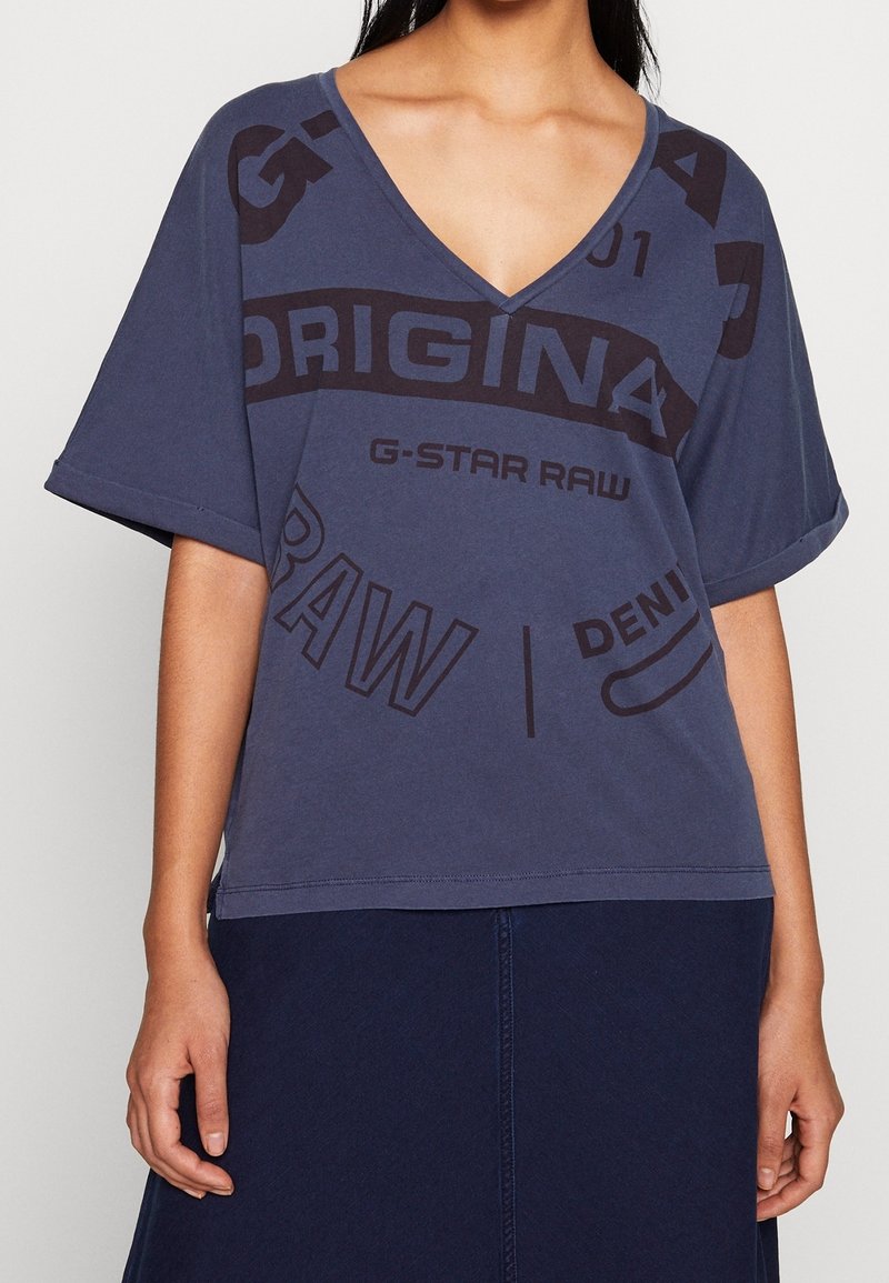 Dunkelblauses, übergroßes T-Shirt mit V-Ausschnitt, das große schwarze Grafiktexte und Logos auf der Vorderseite zeigt. Kurze Ärmel und eine lockere Passform.