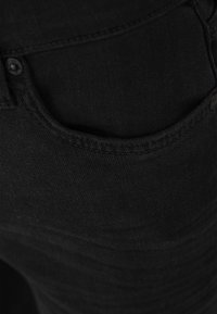 Jeans en denim noir avec une texture lisse, dotés d'une poche avant et de détails en métal sur le bouton, affichant un design ajusté.