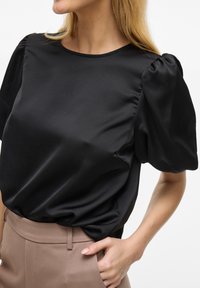 Blusa di raso nera con maniche a palloncino corte, scollatura rotonda e texture liscia, abbinata a pantaloni beige chiaro.