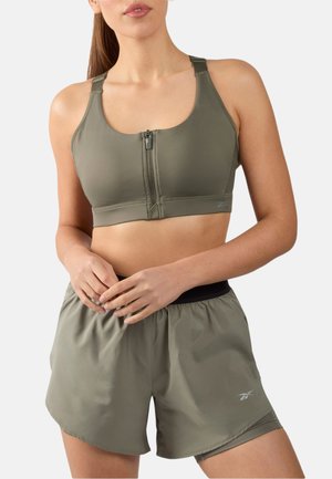 Frau trägt ein olivgrünes Sport-BH mit Reißverschluss vorne und passende Sportshorts, steht mit den Händen in der Taille verschränkt.